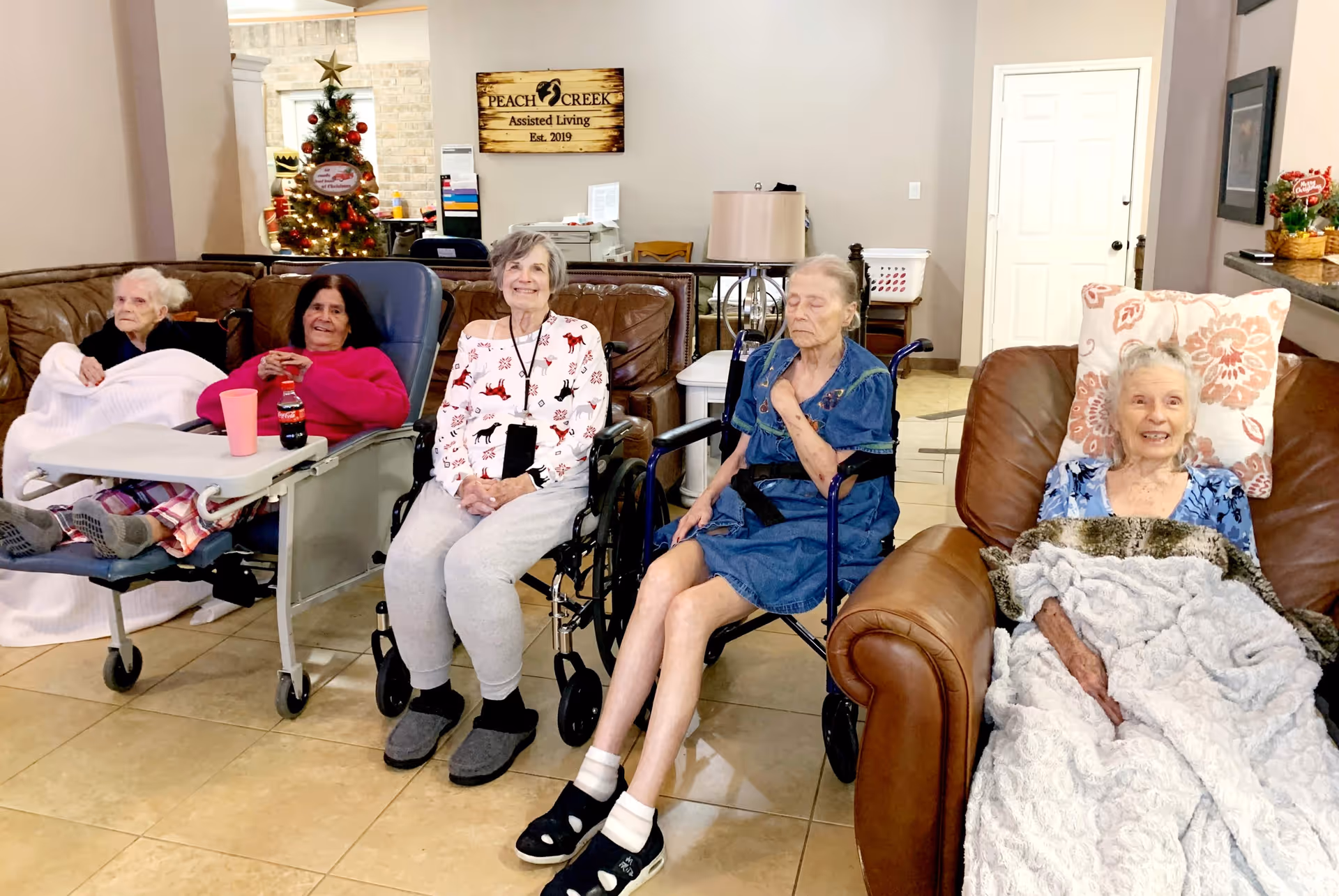 Five elderly residents sitting in chairs and wheelchairs in a communal living room area of Peach Creek Assisted Living, with a holiday tree and facility sign in the background.