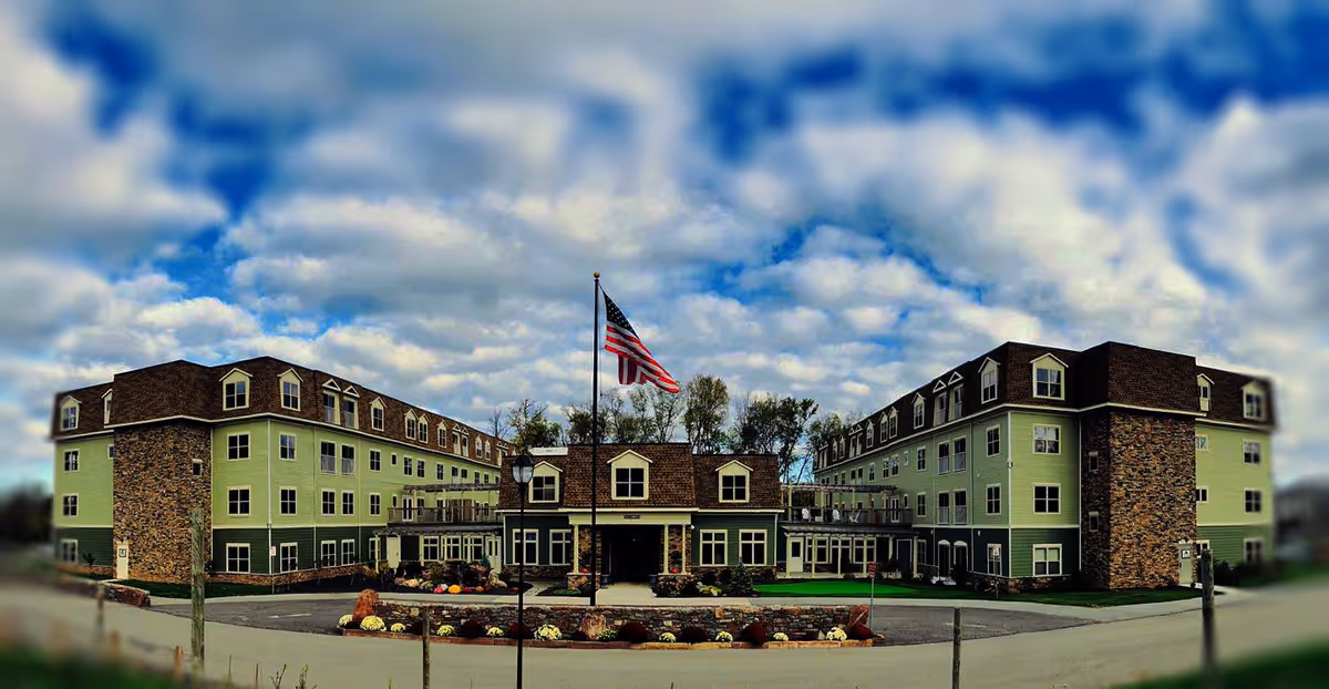 Exterior view of a large senior living facility building with green and stone facade, multiple windows, and an American flag flying on a flagpole in front. The sky is partly cloudy.