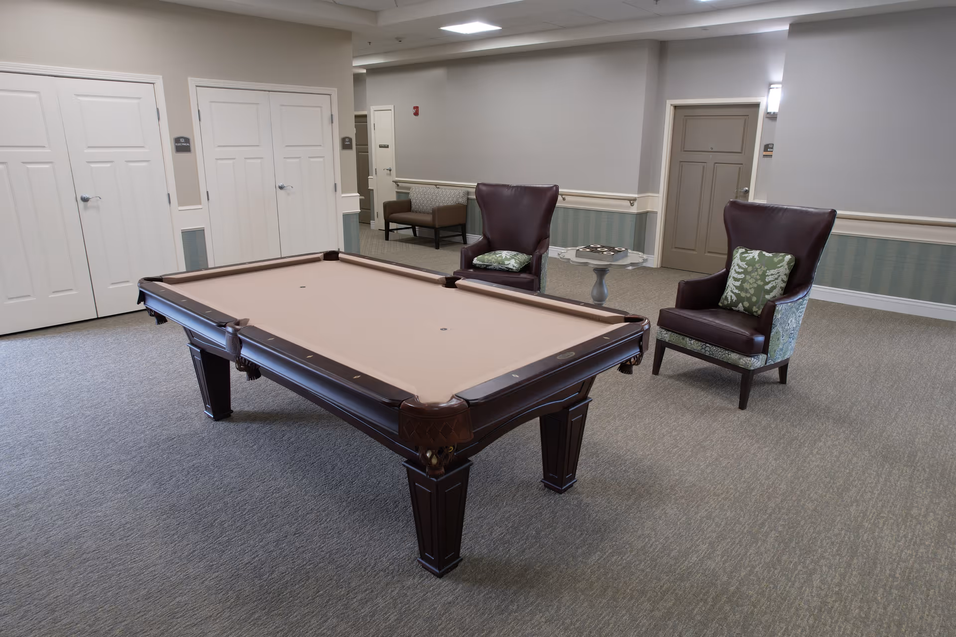 A spacious room with a pool table in the center, two leather armchairs with patterned cushions, a small round table with a board game, and a bench against the wall. The room has beige walls with white trim and multiple closed doors.