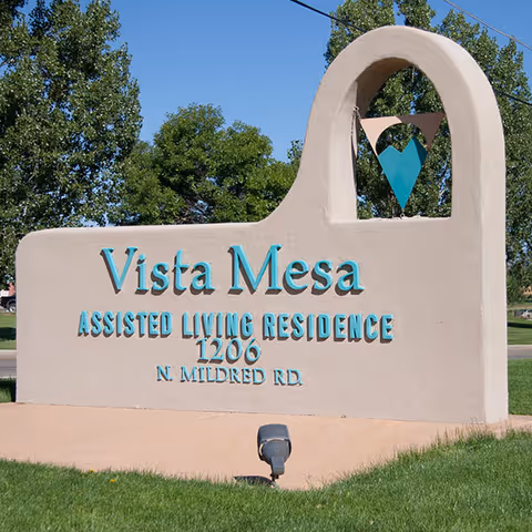 Outdoor sign for Vista Mesa Assisted Living Residence located at 1206 N. Mildred Rd, with a decorative arch and heart-shaped design, surrounded by green grass and trees under a clear blue sky.