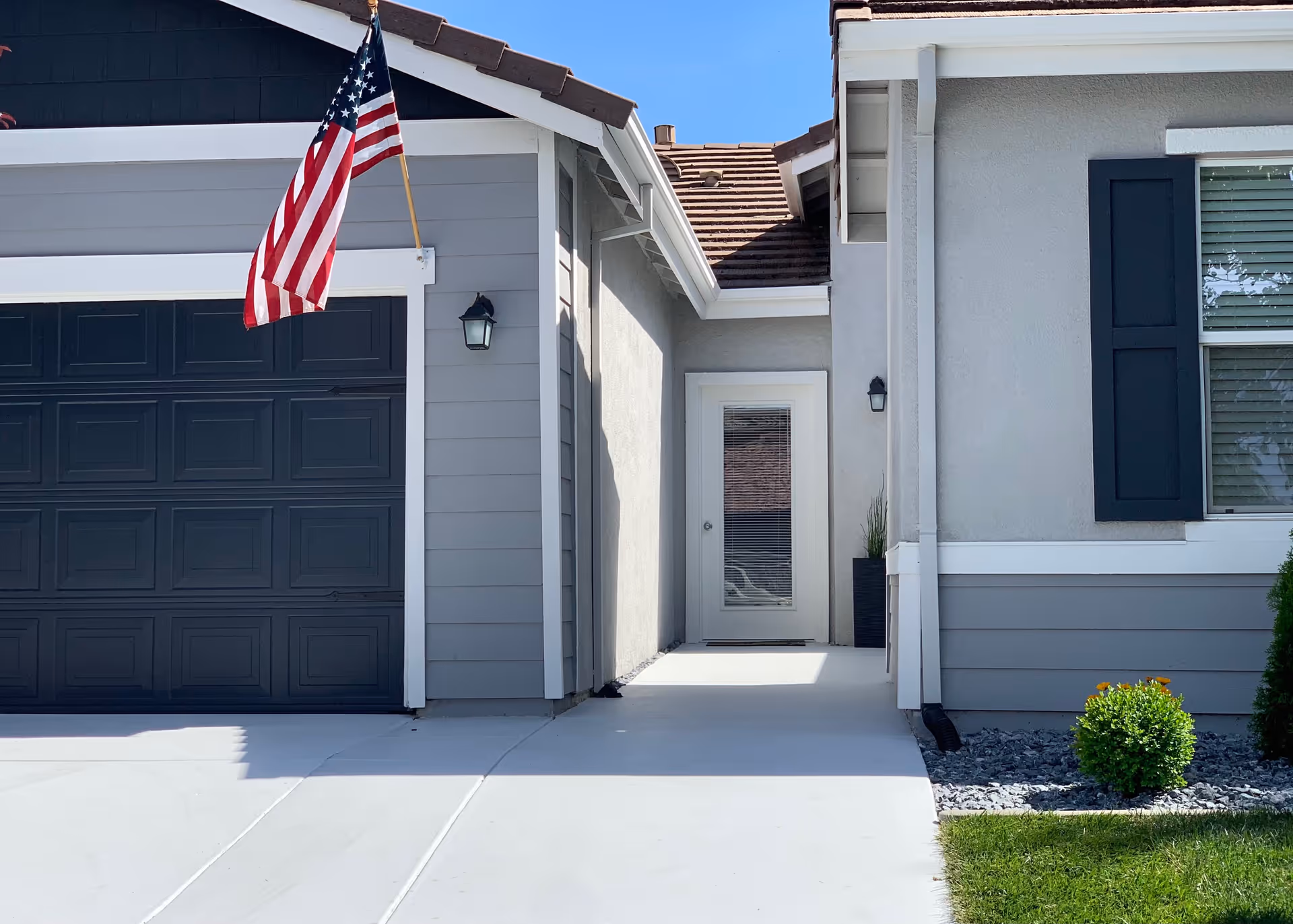 Exterior view of a residential building entrance with a gray garage door, an American flag mounted on the wall, a white door with glass panels, and a small landscaped area with green bushes and rocks.
