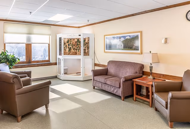 A bright communal sitting room with two leather armchairs, a loveseat, side table and lamp, large windows, and a white enclosed display in the corner.