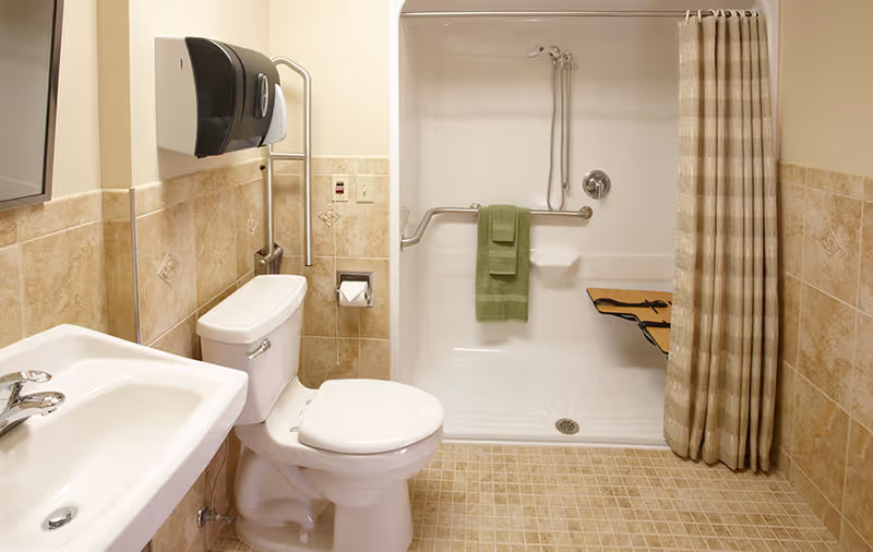 A clean and accessible bathroom featuring a white toilet, a white sink with a chrome faucet, and a walk-in shower with a foldable wooden seat and grab bars. The shower has a beige and white striped curtain and a green towel hanging on the grab bar. The walls are tiled halfway up with beige tiles, and the floor is covered with small square beige tiles. A black paper towel dispenser is mounted on the wall above the toilet.