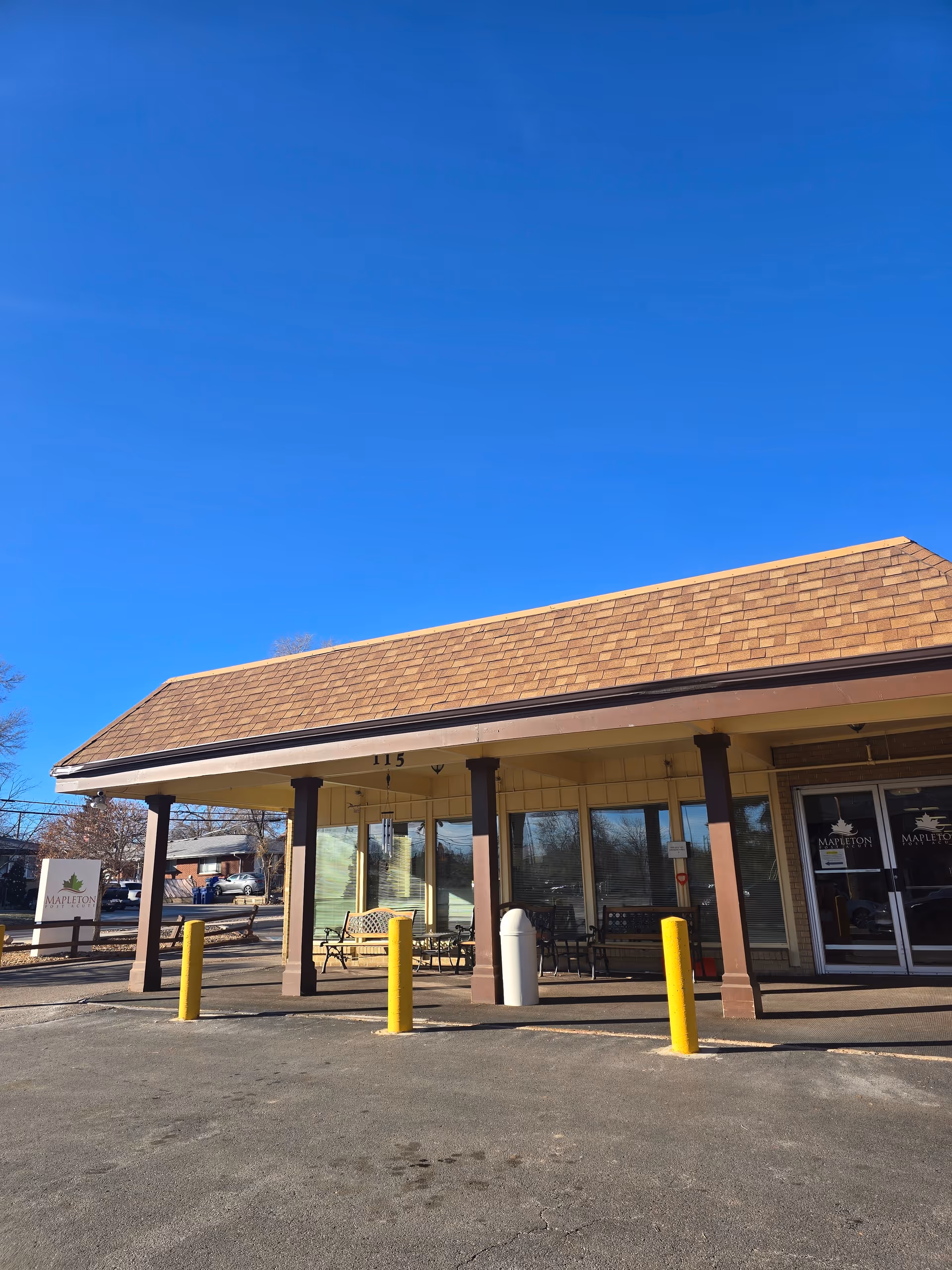 Exterior view of Mapleton Care Center building with a brown shingled roof, beige walls, and large windows. There are yellow bollards in front of the entrance and a sign with the Mapleton Care Center logo on the left side. The sky is clear and blue.