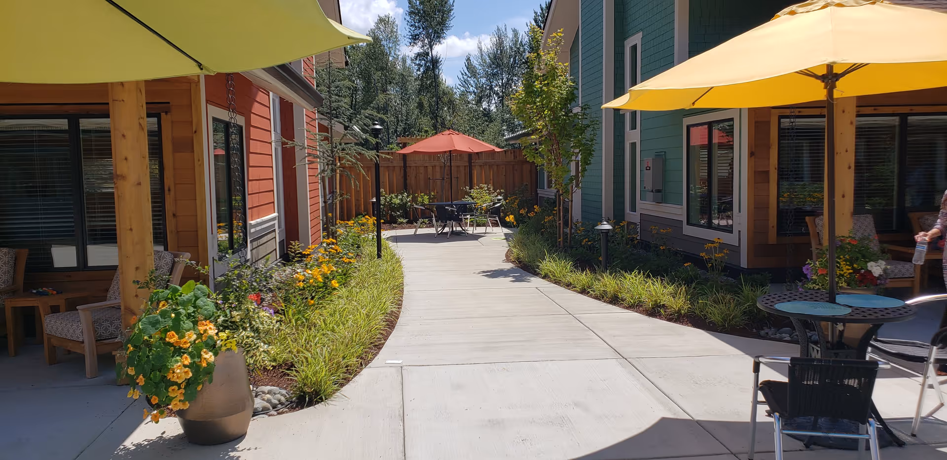 Outdoor courtyard area between two buildings with colorful umbrellas providing shade over tables and chairs. There are flower pots with blooming flowers and landscaped garden beds along the walkway. The buildings are painted in red and green with large windows.