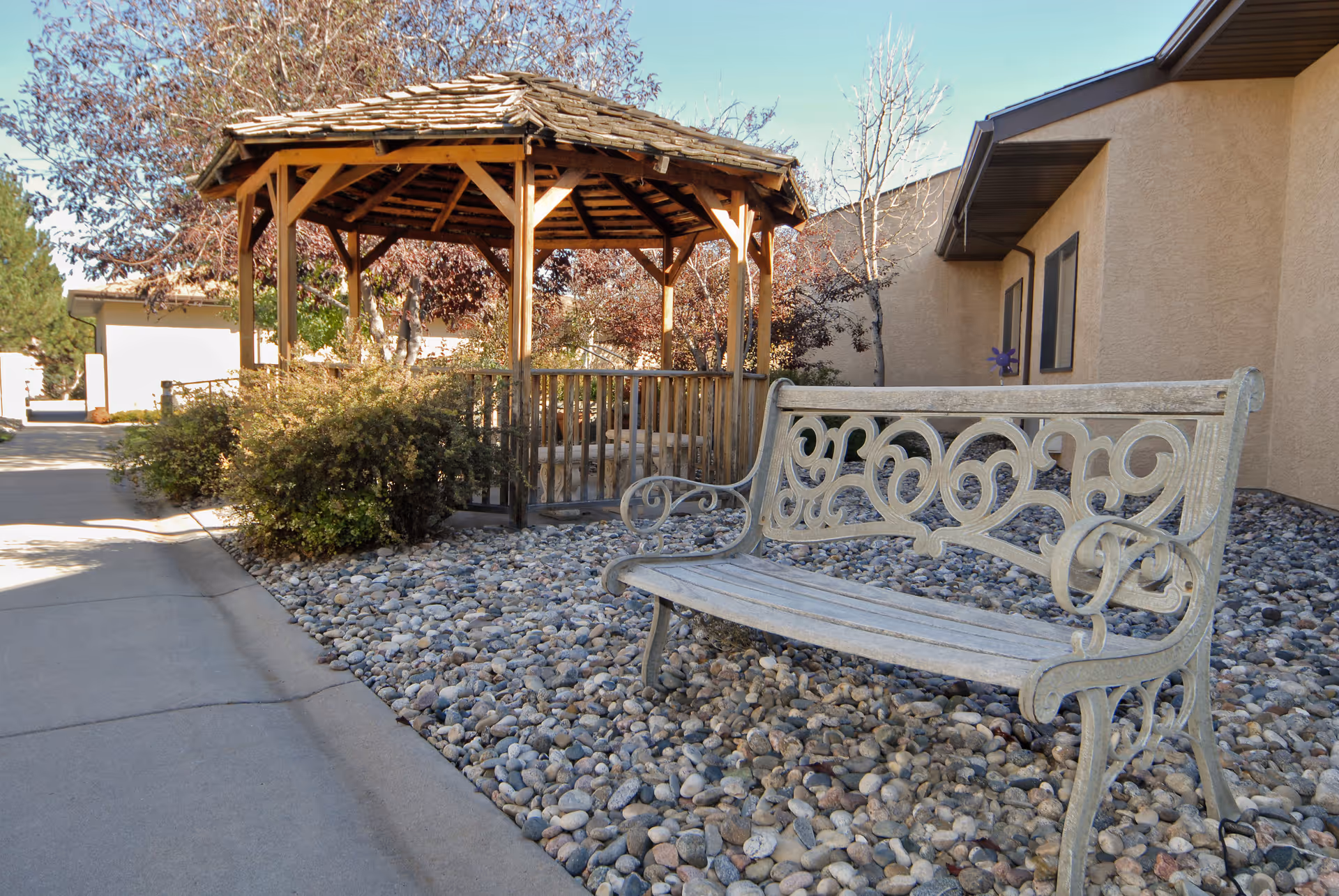 Decorative metal bench on pebbled landscaping beside a wooden gazebo outside a beige building.