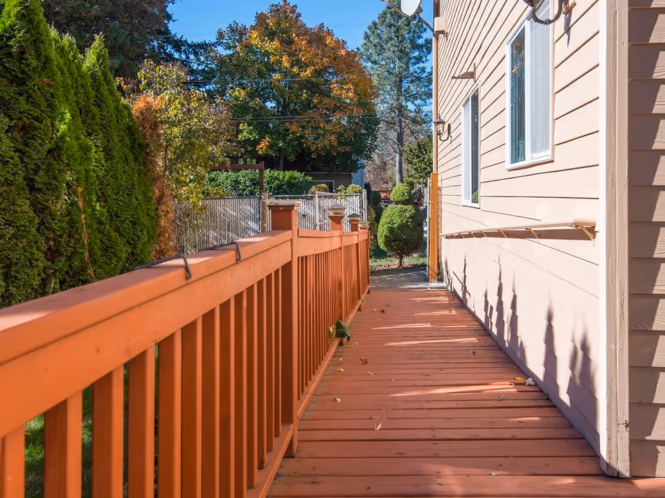 Outdoor wooden ramp with orange railing alongside a beige building, surrounded by green trees and bushes under a clear blue sky.