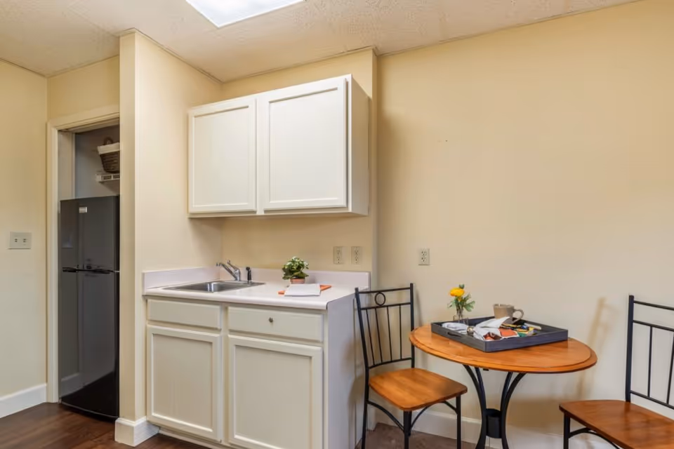 Small kitchenette area with white cabinets, a sink, and a black refrigerator in an open pantry. Next to the kitchenette is a small round wooden table with two chairs, a tray with items, and a small flower vase on the table.
