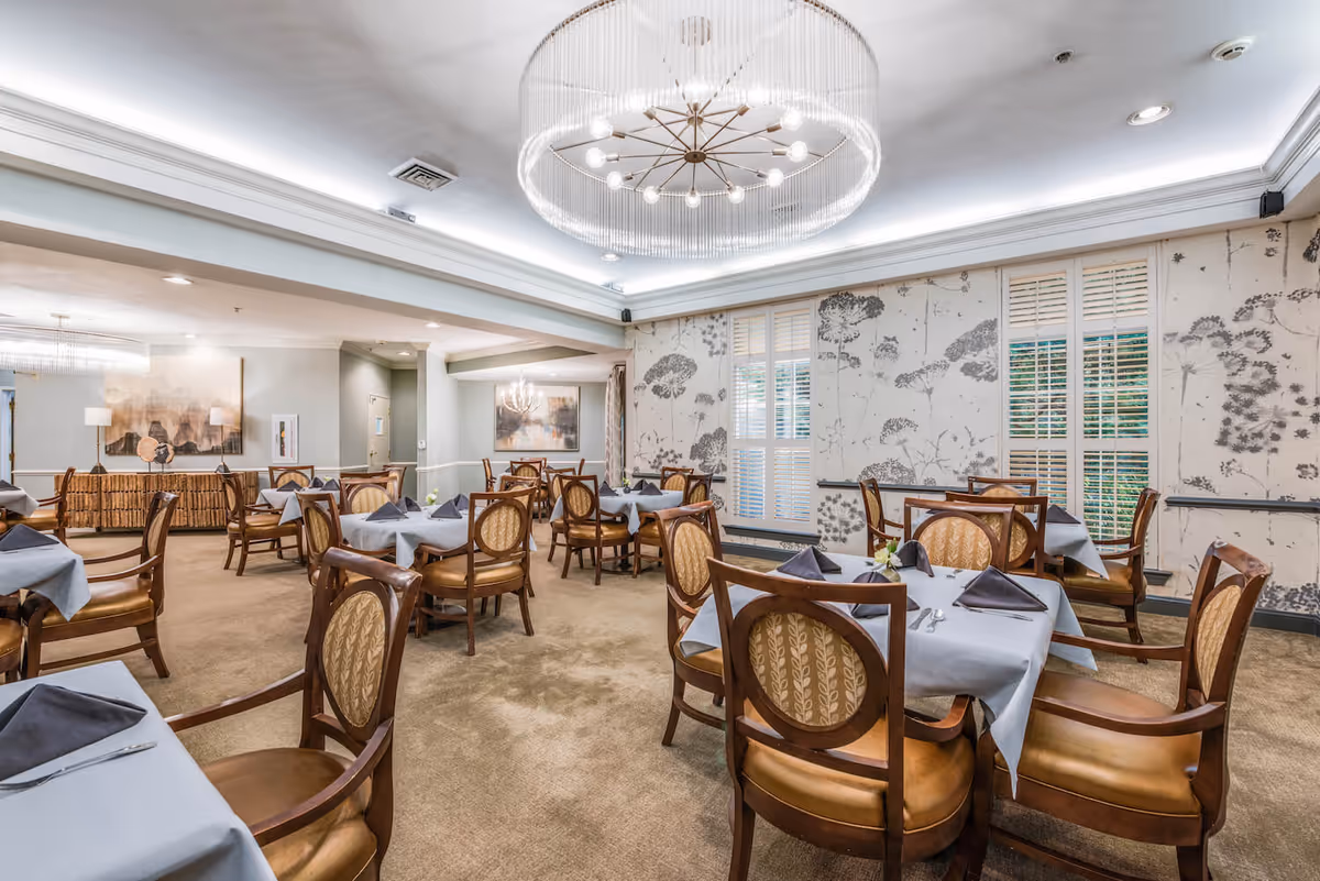 A well-lit dining room with multiple tables covered in light blue tablecloths and set with dark napkins and silverware. The room features elegant wooden chairs with patterned upholstery, large windows with white shutters, floral wallpaper, and modern chandeliers hanging from the ceiling.