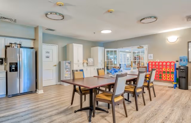 A bright and clean common area in a senior living facility featuring a rectangular table with six chairs around it. In the background, there is a stainless steel refrigerator, white cabinets, a large Connect Four game, and a water dispenser. The room has light-colored walls and wood-style flooring, with glass double doors leading to another room.