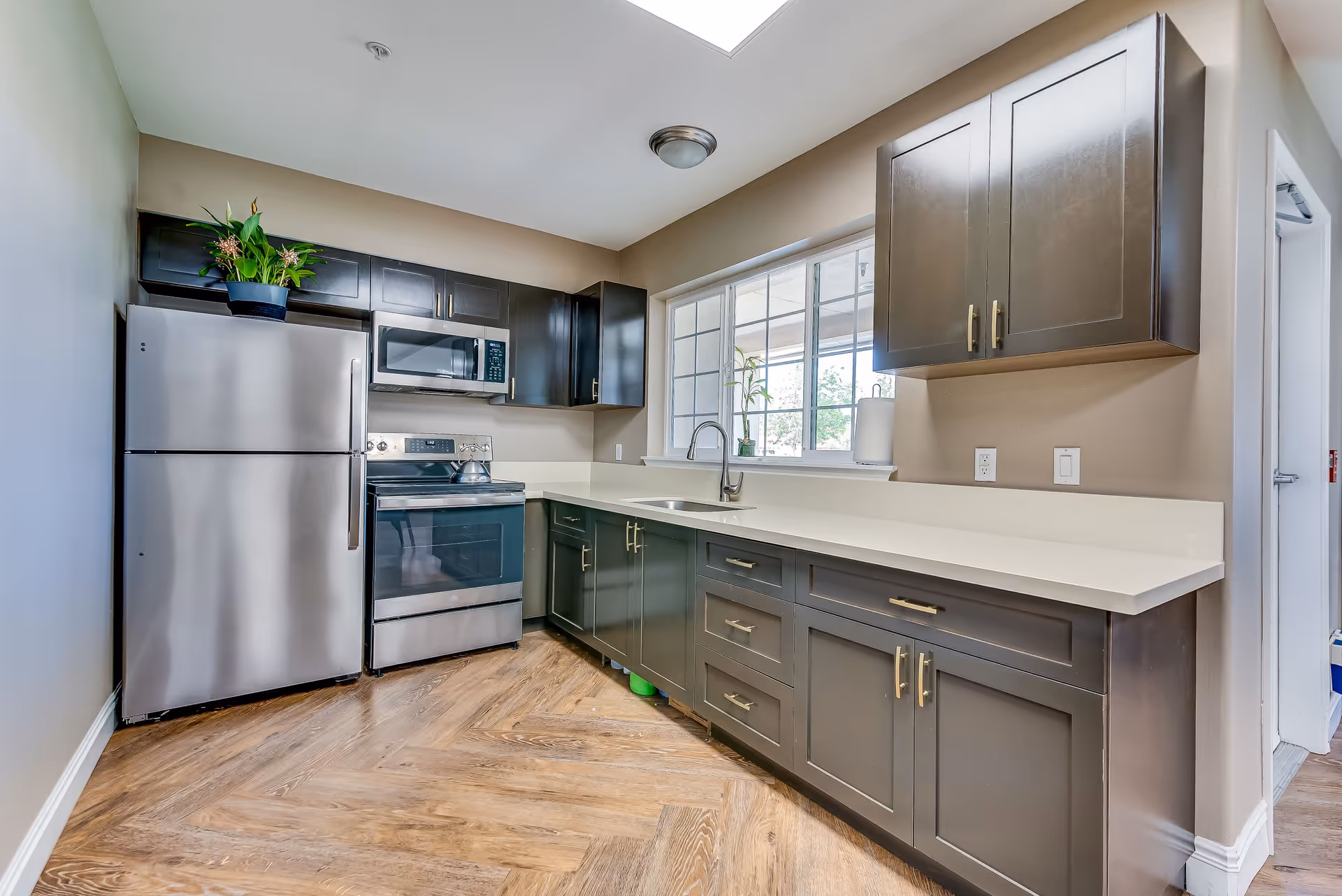Modern kitchen with stainless steel refrigerator, stove, and microwave. Dark gray cabinets with brass handles, white countertops, a sink under a large window, and wood-patterned flooring.