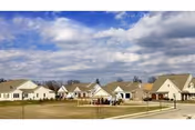 A row of single-story residential buildings with beige siding and brown roofs under a partly cloudy sky, situated along a paved road with a grassy area in front, likely part of a senior living community.