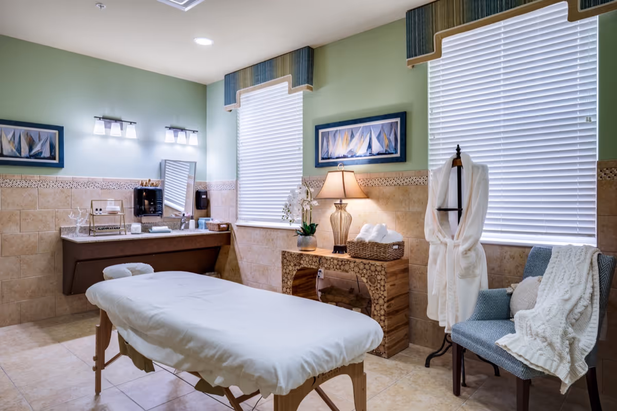Calm spa-like treatment room with a massage table, sink vanity, lamp, robe on a stand, and a chair by two windows.