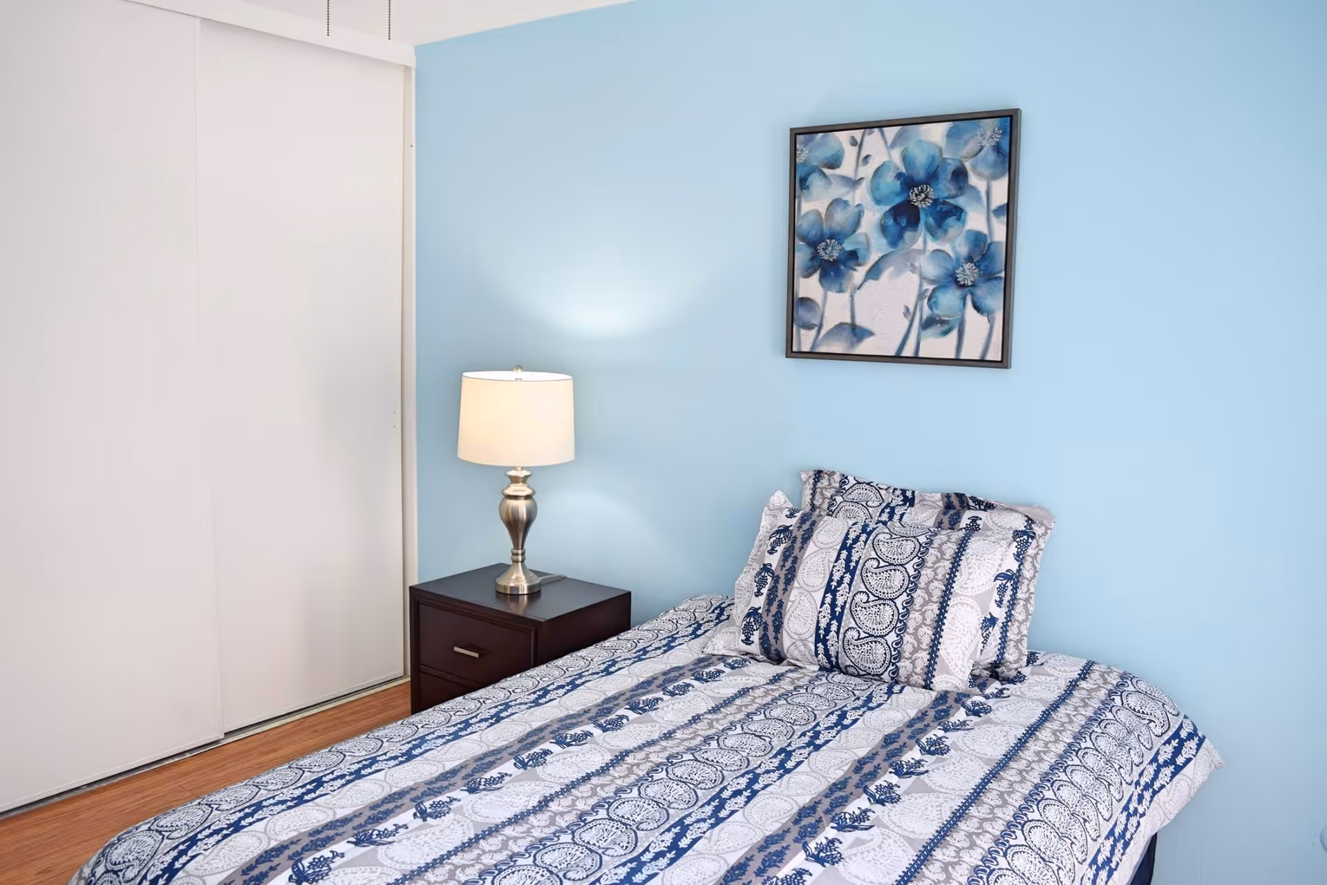 A bedroom with a single bed covered in a blue and white patterned bedspread and matching pillows. Next to the bed is a dark wooden nightstand with a silver lamp that has a white lampshade. The wall behind the bed is painted light blue and features a framed artwork of blue flowers. There is a white sliding closet door on the left side of the room and a wooden floor.