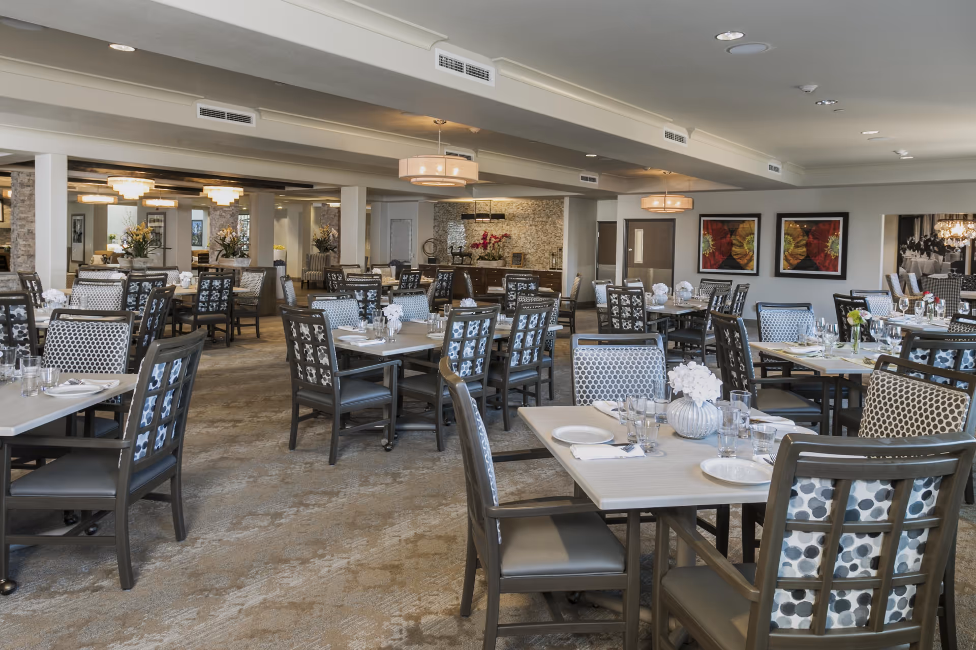 Spacious dining room with multiple tables and chairs arranged neatly. Each table is set with plates, glasses, and napkins, and decorated with small flower arrangements. The room features modern lighting fixtures, carpeted floors, and framed artwork on the walls.