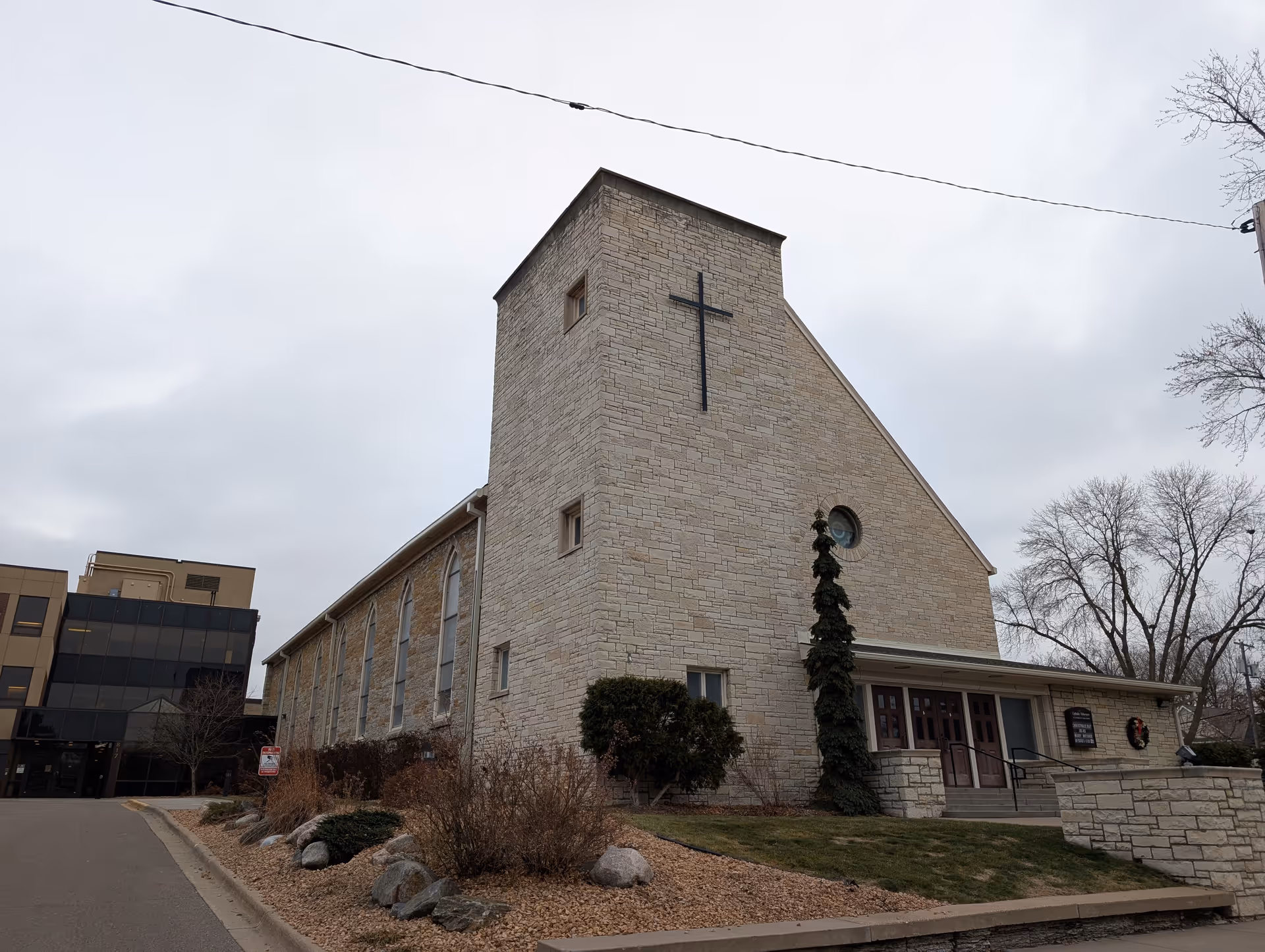 Exterior view of a stone building with a large cross mounted on the front tower, adjacent to a modern multi-story building. The scene includes a small landscaped area with bushes and rocks, and a cloudy sky overhead.