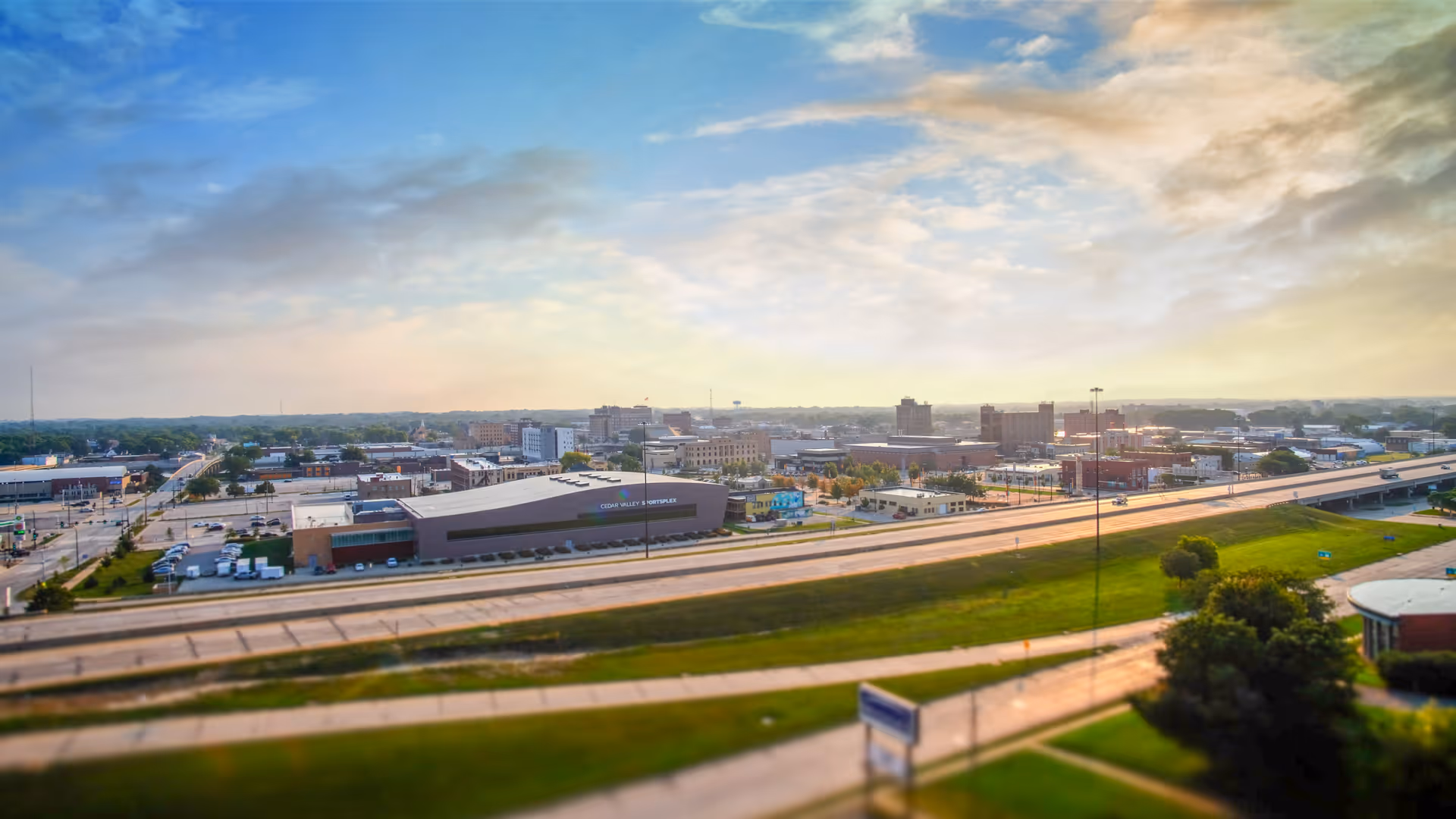 A panoramic view of a cityscape with a highway in the foreground, various buildings, and a partly cloudy sky during sunset or sunrise.
