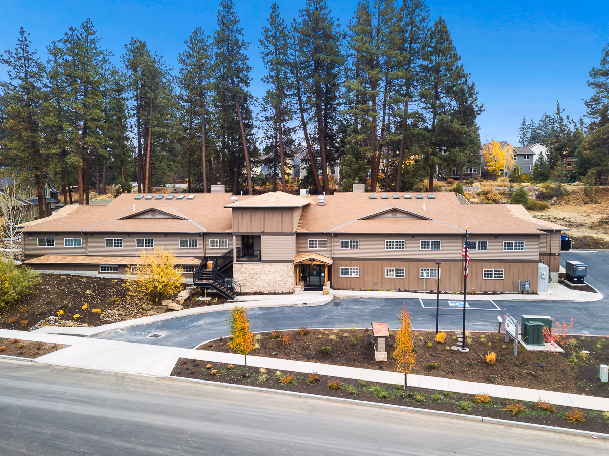 Exterior front view of a two-story brown building surrounded by trees and landscaping with a driveway and parking area in front. The building has multiple windows, a central entrance with a small covered porch, and an American flag on a flagpole near the entrance.