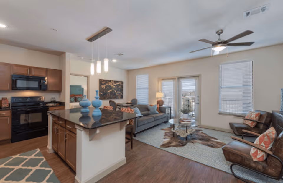 A modern open-concept living space featuring a kitchen with wooden cabinets, black appliances, and a large island with a black countertop and blue decorative vases. The living room area includes a gray sofa with colorful pillows, two brown leather chairs with patterned cushions, a glass coffee table on a rug, and a ceiling fan. Large windows and glass doors allow natural light into the room.