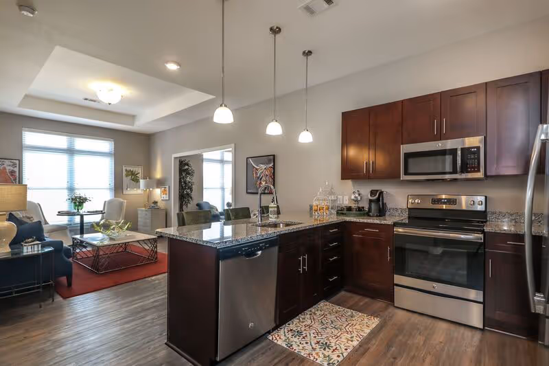 Modern kitchen and living room area in a senior living facility. The kitchen features dark wood cabinets, stainless steel appliances including a microwave, oven, dishwasher, and refrigerator. A granite countertop island with a sink and three pendant lights hangs above. The living room has a large window with blinds, a red area rug, a coffee table, chairs, and a sofa. The space is well-lit with natural and artificial light.