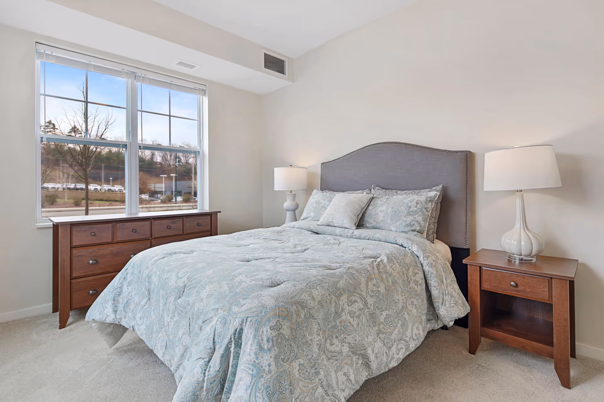 A bedroom with a large bed covered in a light blue patterned comforter and matching pillows. The bed has a gray upholstered headboard. On either side of the bed are wooden nightstands with white lamps. A wooden dresser is positioned under a large window showing an outdoor view with trees and parked cars.