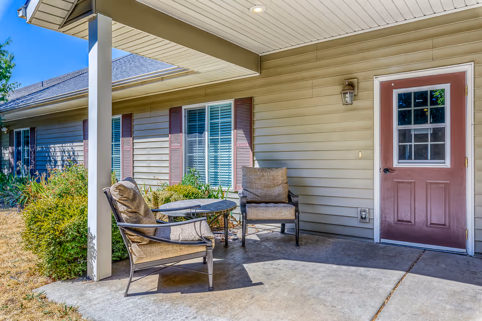 Covered porch area with two cushioned chairs and a small round table beside a red entry door on a single-story building.