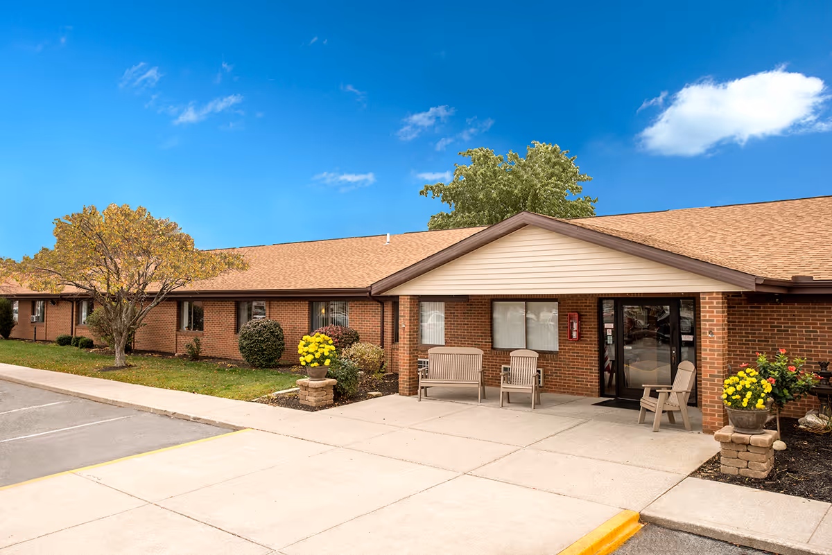 Exterior view of Grande Lake Healthcare Center showing a single-story brick building with a brown shingled roof. There are three chairs placed on the concrete porch near the entrance door. The area is landscaped with green bushes, yellow flowers in planters, and a tree with autumn-colored leaves. The sky is clear with a few clouds.