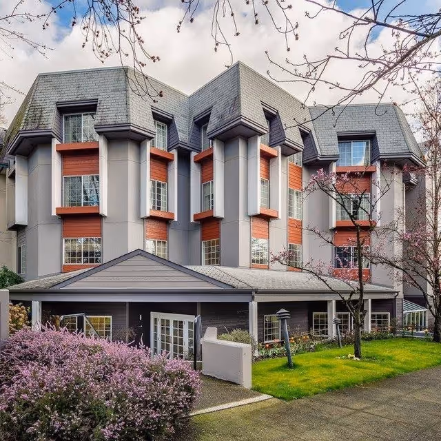 Exterior view of a multi-story residential building with gray and red accents, surrounded by blooming trees and green grass under a partly cloudy sky.
