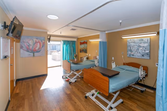 A room with two hospital-style beds with blue bedding, each equipped with a bedside table and medical equipment. The room has wooden flooring, beige walls, and two paintings on the walls. A large window with turquoise curtains lets in natural light. A wall-mounted TV is visible on the left side.