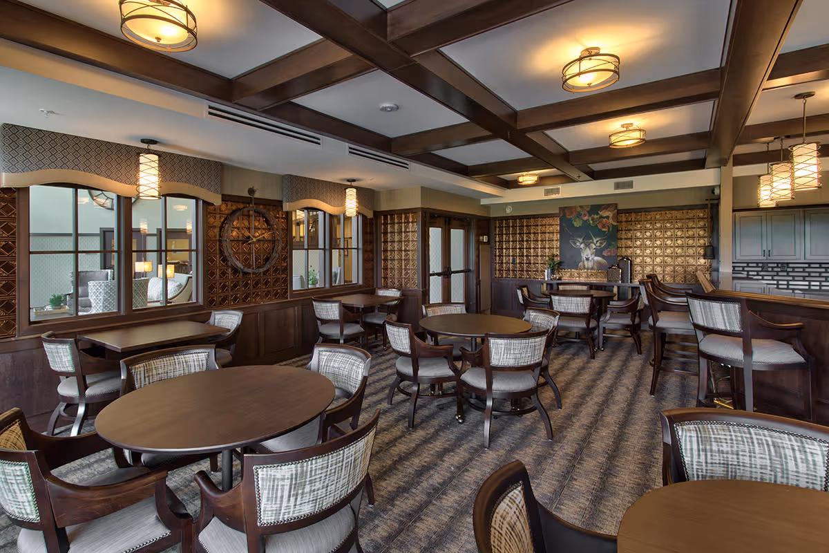 A cozy dining room with multiple round and square wooden tables surrounded by cushioned chairs. The room features a patterned carpet, wooden ceiling beams, and warm lighting fixtures. Decorative wall panels and a painting of a deer with flowers add to the ambiance. A bar area with high chairs is visible on the right side.