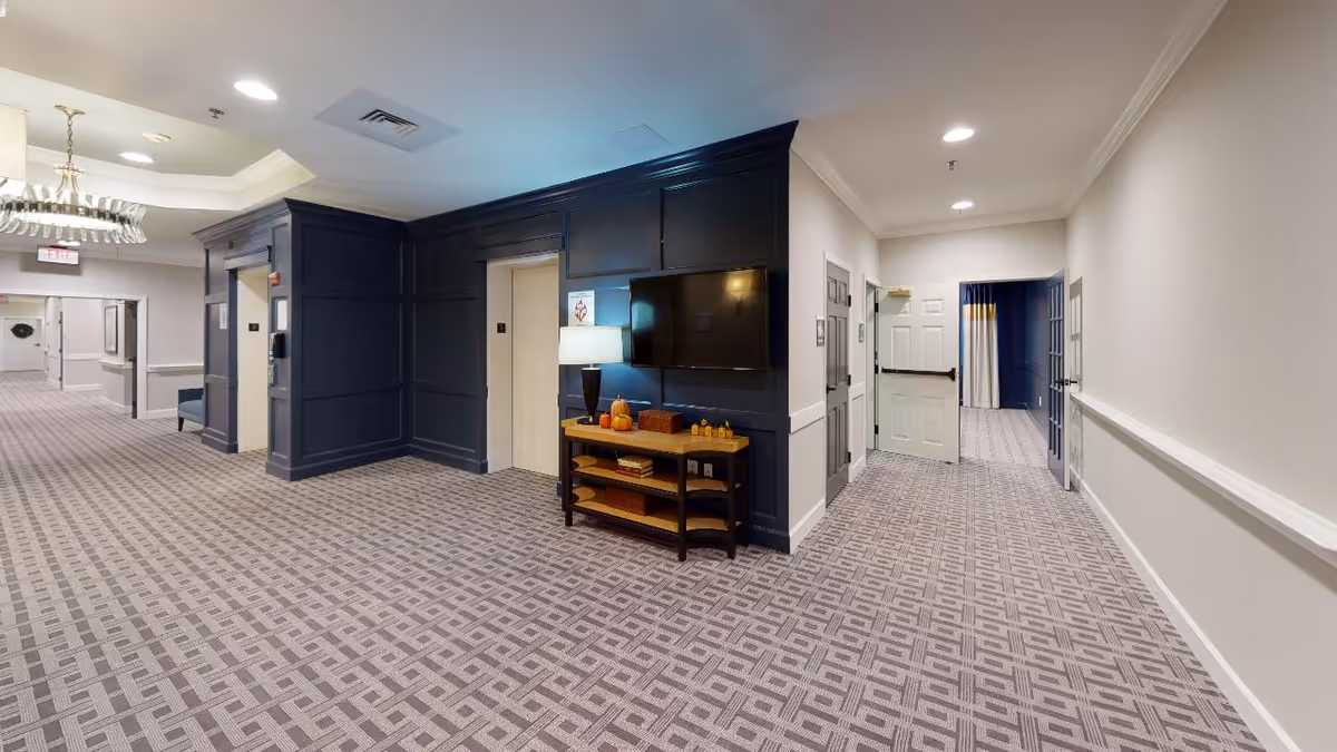 Carpeted interior hallway and common area with elevators, a wall-mounted TV on dark paneling and a small console table with a lamp and decor.