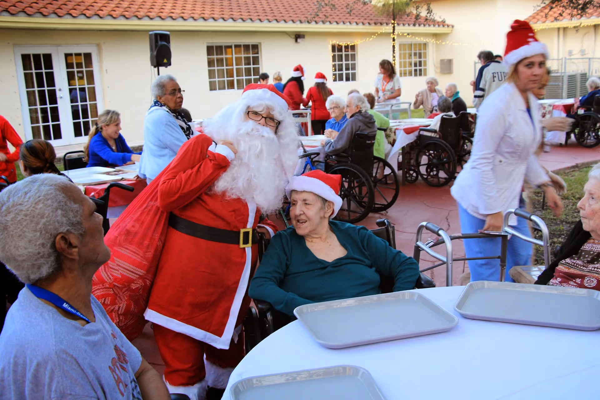 A festive outdoor gathering at an assisted living facility with elderly residents, some in wheelchairs, seated around tables with trays. A person dressed as Santa Claus interacts with a resident wearing a Santa hat. Other staff and residents are visible in the background, some wearing holiday hats, enjoying the event.
