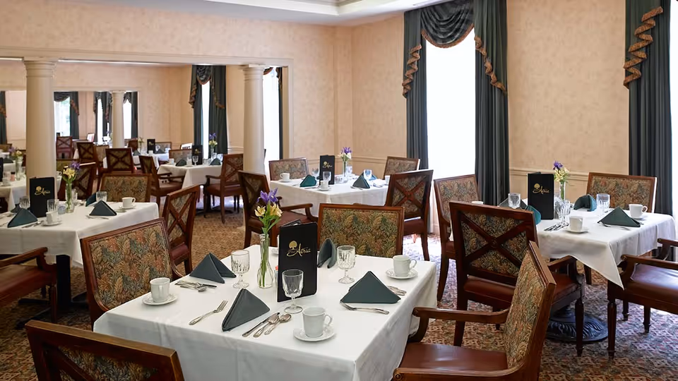 A dining room with several tables set for a meal, each covered with white tablecloths, dark green folded napkins, cups, glasses, and silverware. The chairs have wooden frames with patterned upholstery. There are vases with flowers on the tables, and large windows with dark green curtains letting in natural light. The room has beige walls and carpeted floors with a decorative pattern.