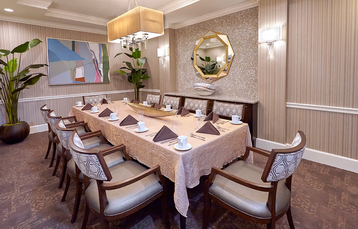 A formal dining room with a long rectangular table covered with a beige tablecloth, set with white cups, saucers, glasses, silverware, and brown folded napkins. The room features patterned upholstered chairs, a decorative wall mirror, two large potted plants, a modern chandelier, and abstract wall art.
