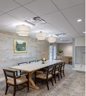 A spacious dining room with a long wooden table surrounded by ten chairs. The room features a light-colored wood accent wall with a framed artwork, three large modern pendant lights hanging from the ceiling, and a patterned carpet floor. In the background, there is a counter with cabinets and a door leading to another area.