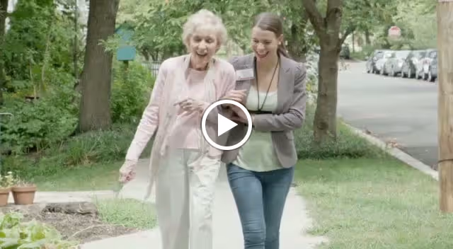 An elderly woman and a younger woman walking arm in arm on a sidewalk in a green, tree-lined neighborhood. Both are smiling and appear happy, with parked cars visible along the street.