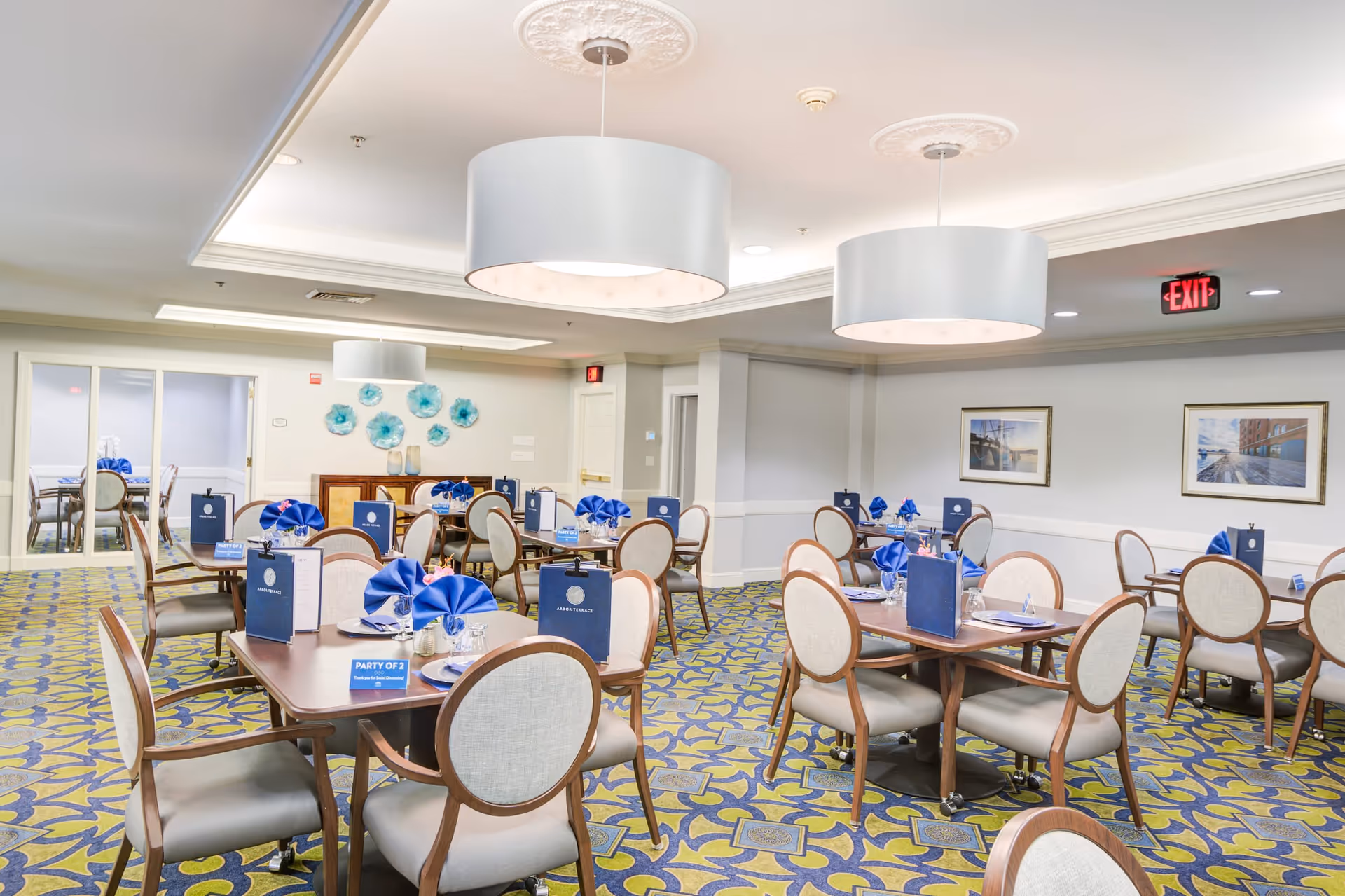 A bright and spacious dining room in a senior living facility with multiple tables set with blue napkins, menus, and place settings. The room features patterned carpet, light-colored walls, framed artwork, and modern pendant lights hanging from the ceiling.