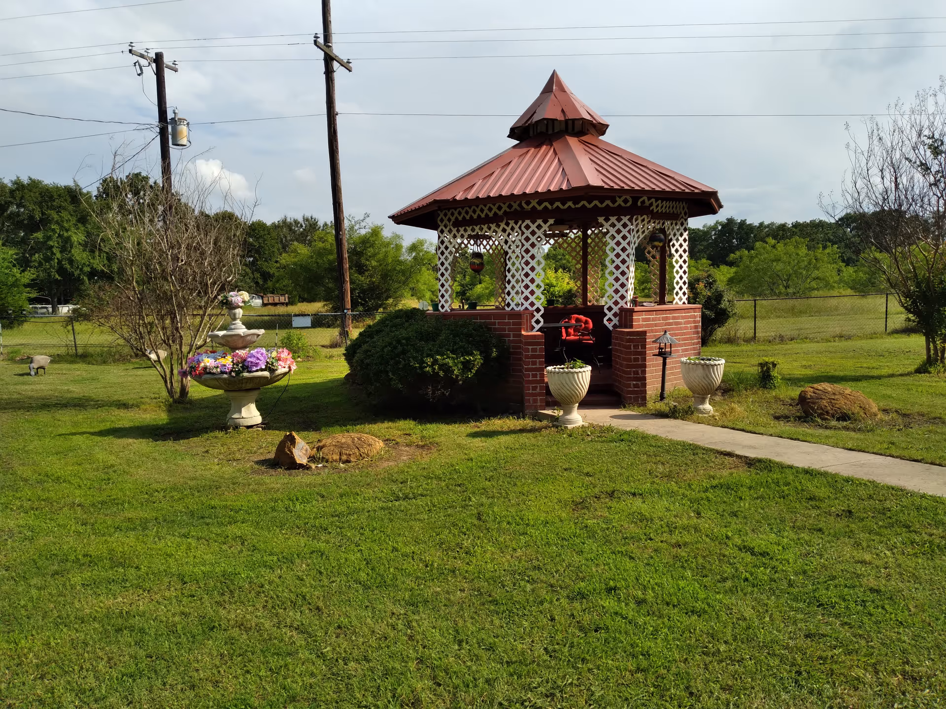 A small outdoor gazebo with a red metal roof and white lattice walls, surrounded by green grass and bushes. There is a concrete pathway leading to the gazebo entrance, two large white planters on either side of the entrance, and a decorative stone fountain with colorful flowers on the left side. The background shows trees, a chain-link fence, and two utility poles with power lines.
