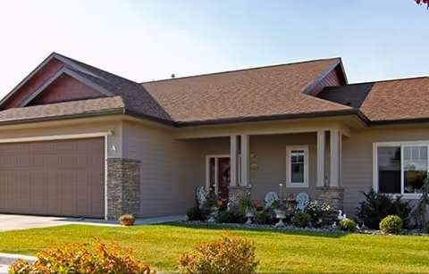 Exterior view of a single-story residential building with a two-car garage, a front porch with columns, and a well-maintained lawn with shrubs and plants.