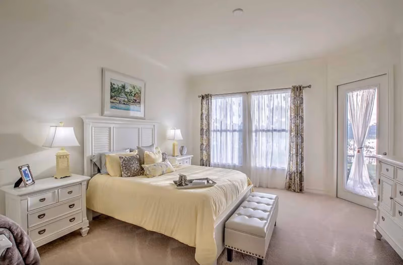 Bright, tastefully decorated bedroom with a large bed, nightstands and lamps, sheer-curtained windows and a door to a balcony.