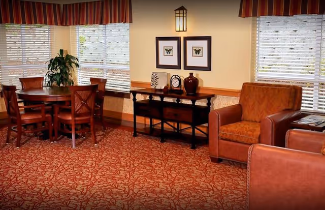 A cozy interior room with a round wooden table surrounded by four chairs on the left, a wooden console table with decorative items and two framed butterfly pictures on the wall above it in the center, and two brown upholstered armchairs on the right. The room has large windows with blinds and red patterned carpet flooring.