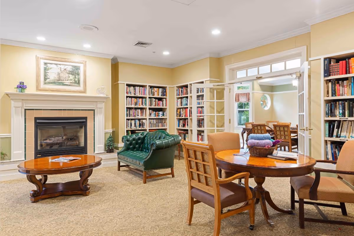 A cozy senior living facility common area with a green leather armchair, a wooden coffee table, and a round wooden table with chairs. The room features built-in bookshelves filled with books, a fireplace with a framed painting above it, and an adjoining room visible through glass double doors.