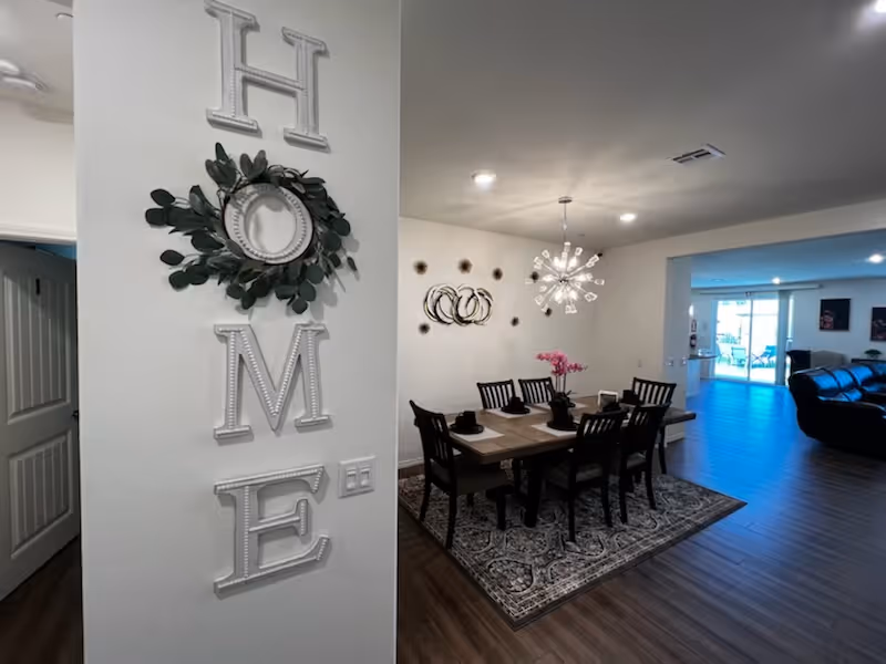 Interior view of a dining area in a care home with a wooden dining table set for six people, a decorative wall with the word HOME vertically arranged with a wreath on the letter O, modern chandelier above the table, and a living room visible in the background with leather sofas and large windows.