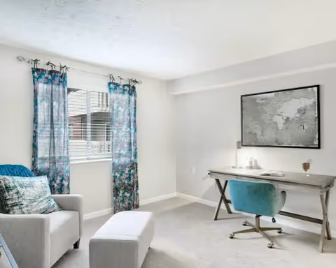 A bright room with a window covered by blue patterned curtains, a gray armchair with a decorative pillow, a matching ottoman, and a wooden desk with a teal swivel chair. A world map is framed and hung on the wall above the desk.