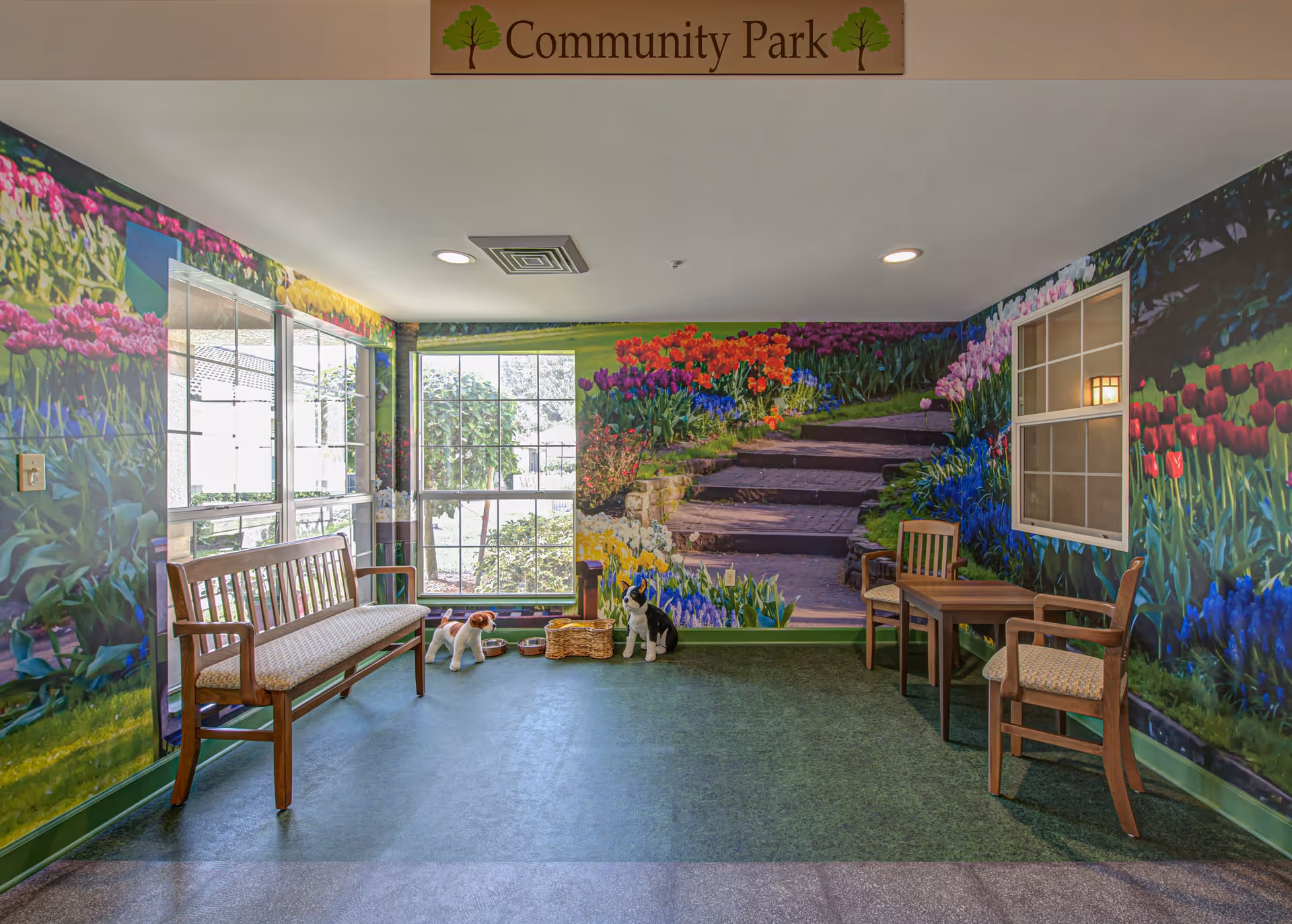An indoor seating area with wooden benches and chairs arranged around a small table. The walls are decorated with a colorful mural depicting a garden scene with flowers and stone steps. There are two large windows letting in natural light, and two dog statues with bowls placed near the wall. A sign above reads 'Community Park'.