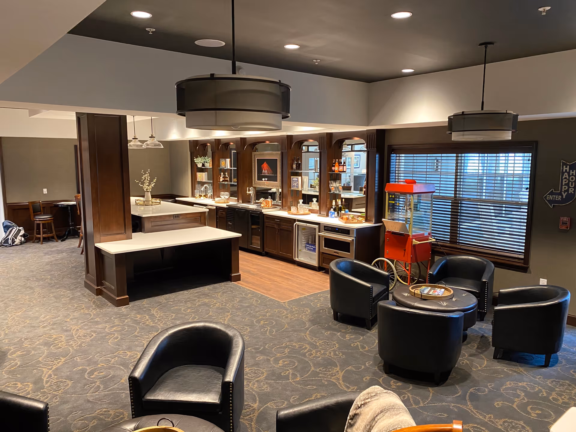 A cozy common area with a small bar setup featuring dark wood cabinets, a countertop with glasses and bottles, and a popcorn machine near a window with blinds. The room has several black leather chairs arranged around a circular ottoman, carpeted flooring with a patterned design, and modern ceiling lights.
