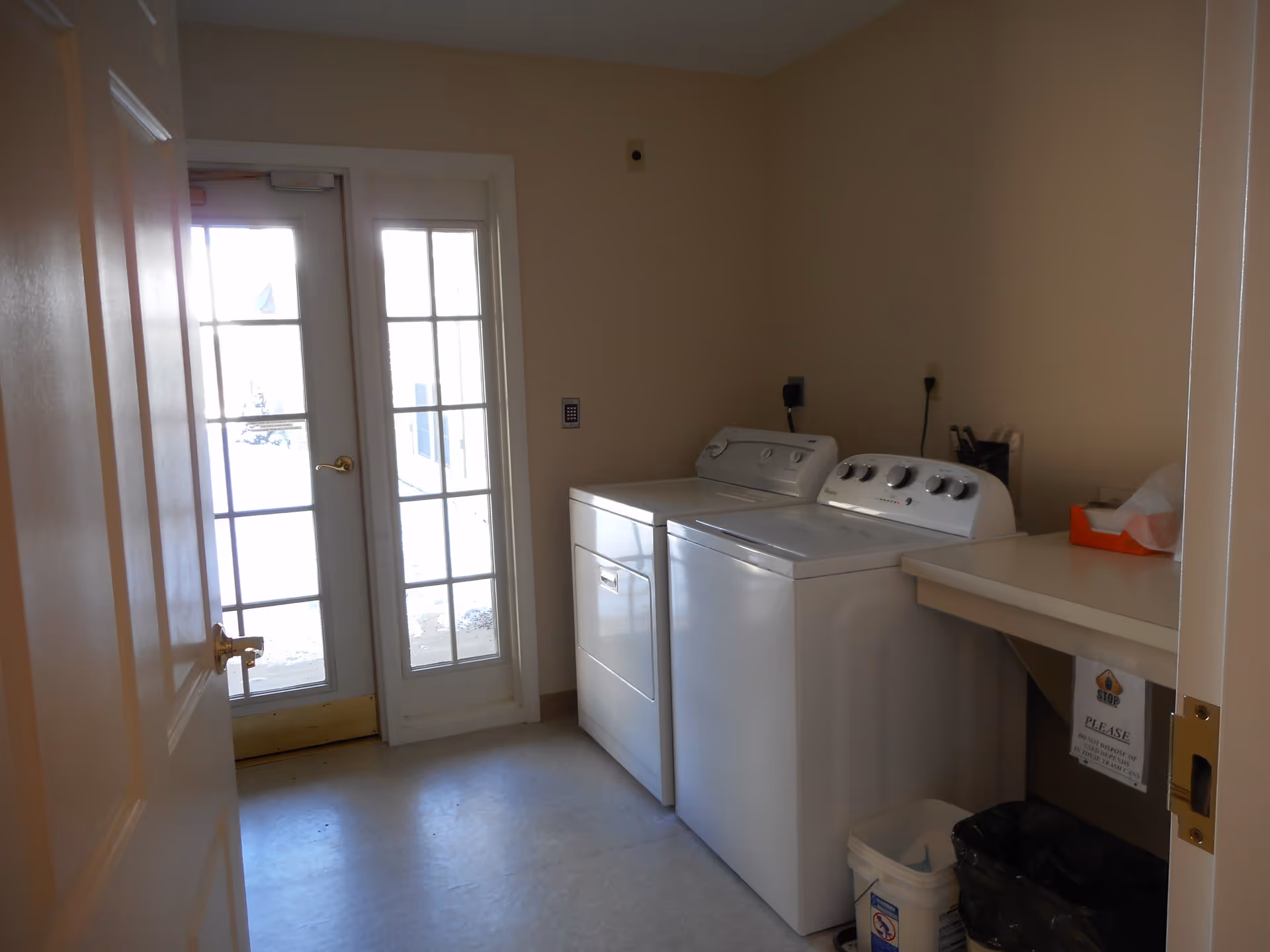 Laundry room with a white washing machine and dryer side by side, a countertop with a tissue box, a trash can, and a door with glass panels letting in natural light.