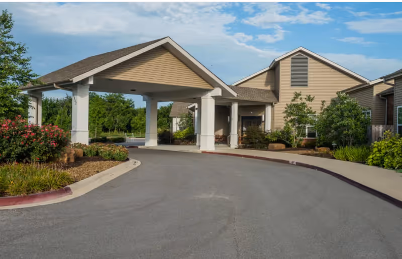 Exterior view of Concord Place Memory Care facility showing a covered driveway entrance with beige siding buildings, landscaped bushes, trees, and a clear blue sky with some clouds.