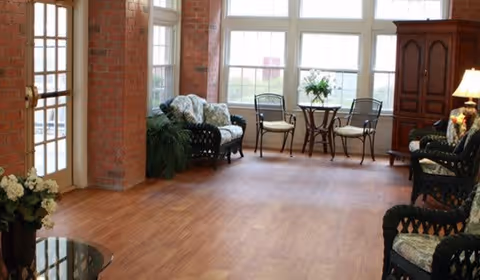 A bright and spacious sitting area with large windows letting in natural light. The room features wooden flooring, a small round table with two metal chairs, a cushioned wicker loveseat, two cushioned wicker armchairs, a wooden cabinet, and decorative plants and flowers. The walls are a mix of exposed brick and white, and there is a glass-paned door on the left side.
