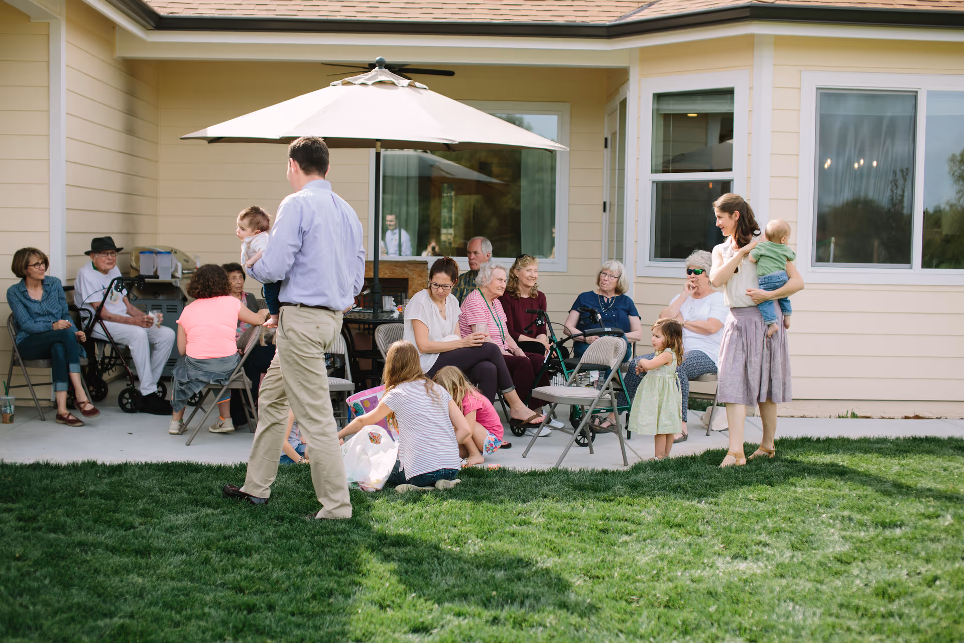 A group of elderly people and children gathered outside a light yellow building with large windows. Some elderly individuals are seated on folding chairs under a large patio umbrella on a concrete patio, while children and adults stand or sit on the grass nearby. The scene appears to be a social gathering in a senior living facility's outdoor area.
